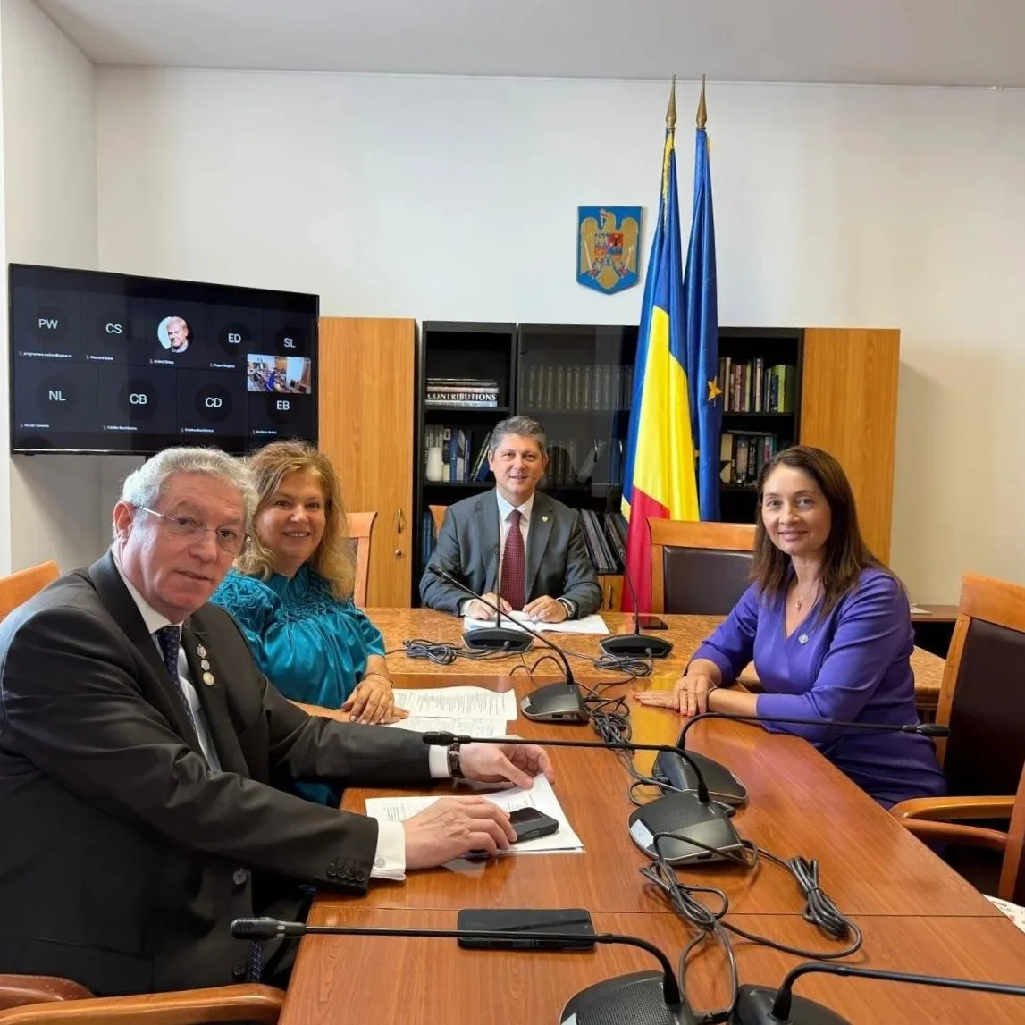 Titus Corlatean  and Four people seated around a conference table with microphones, in a room with flags and a crest, participating in a video call on a screen.