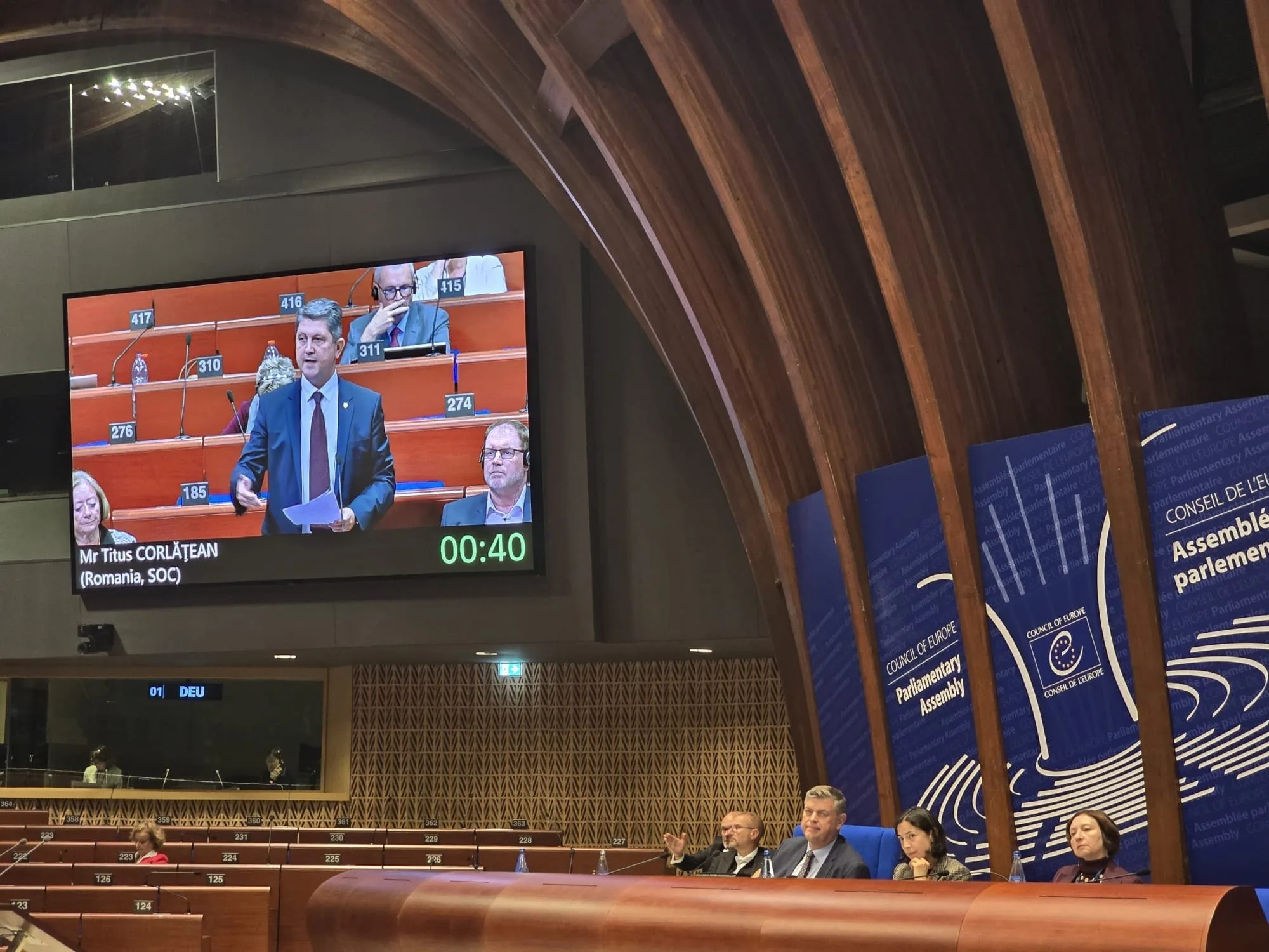 A man in a suit and tie speaking at a parliamentary assembly, with a large screen showing his image and a timer, and three other people seated at a panel table.