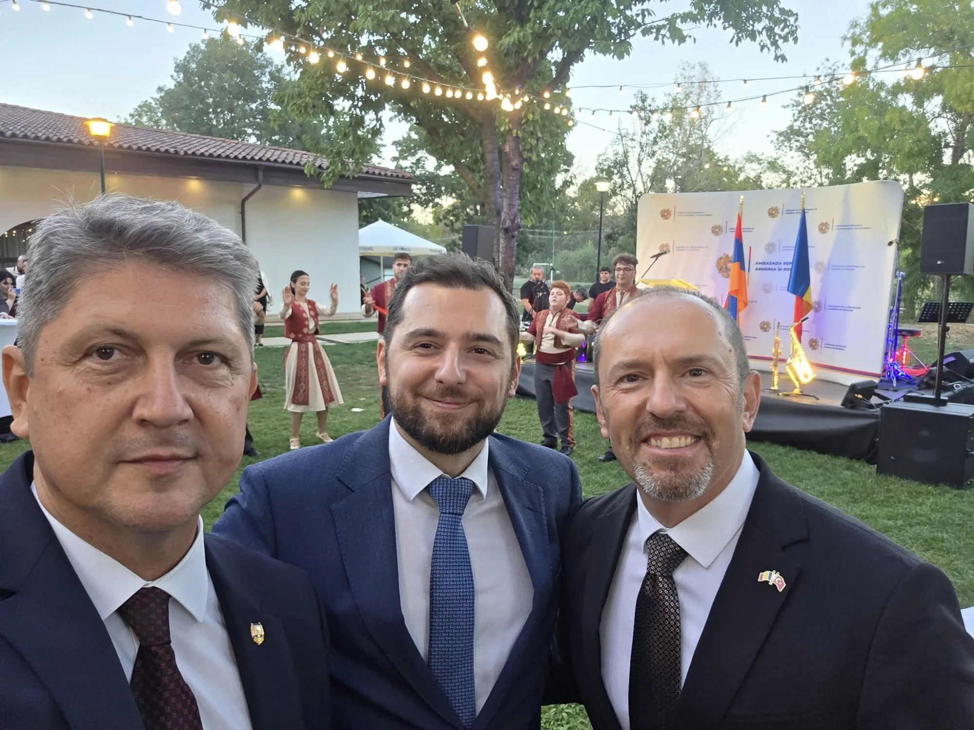 Three men dressed in suits taking a selfie at an outdoor event with a stage, flags, and traditional dancers. String lights are hanging above, and trees are in the background.