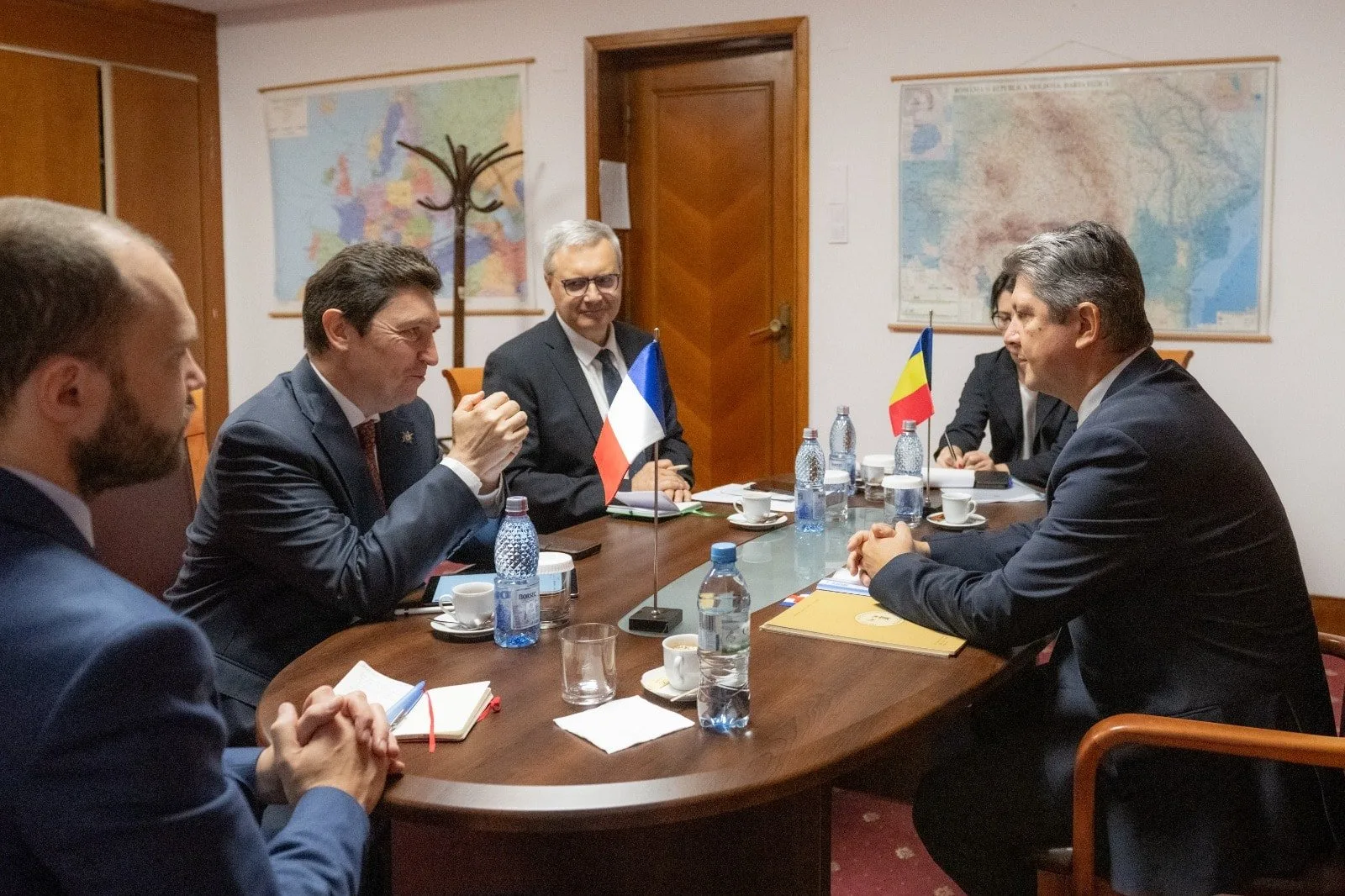 A business meeting with five people sitting around a wooden table in a conference room. Two are holding small flags of France and Romania. The table has bottled water, cups, notebooks, and a folder. Maps of France and Romania are hanging on the white walls behind them.