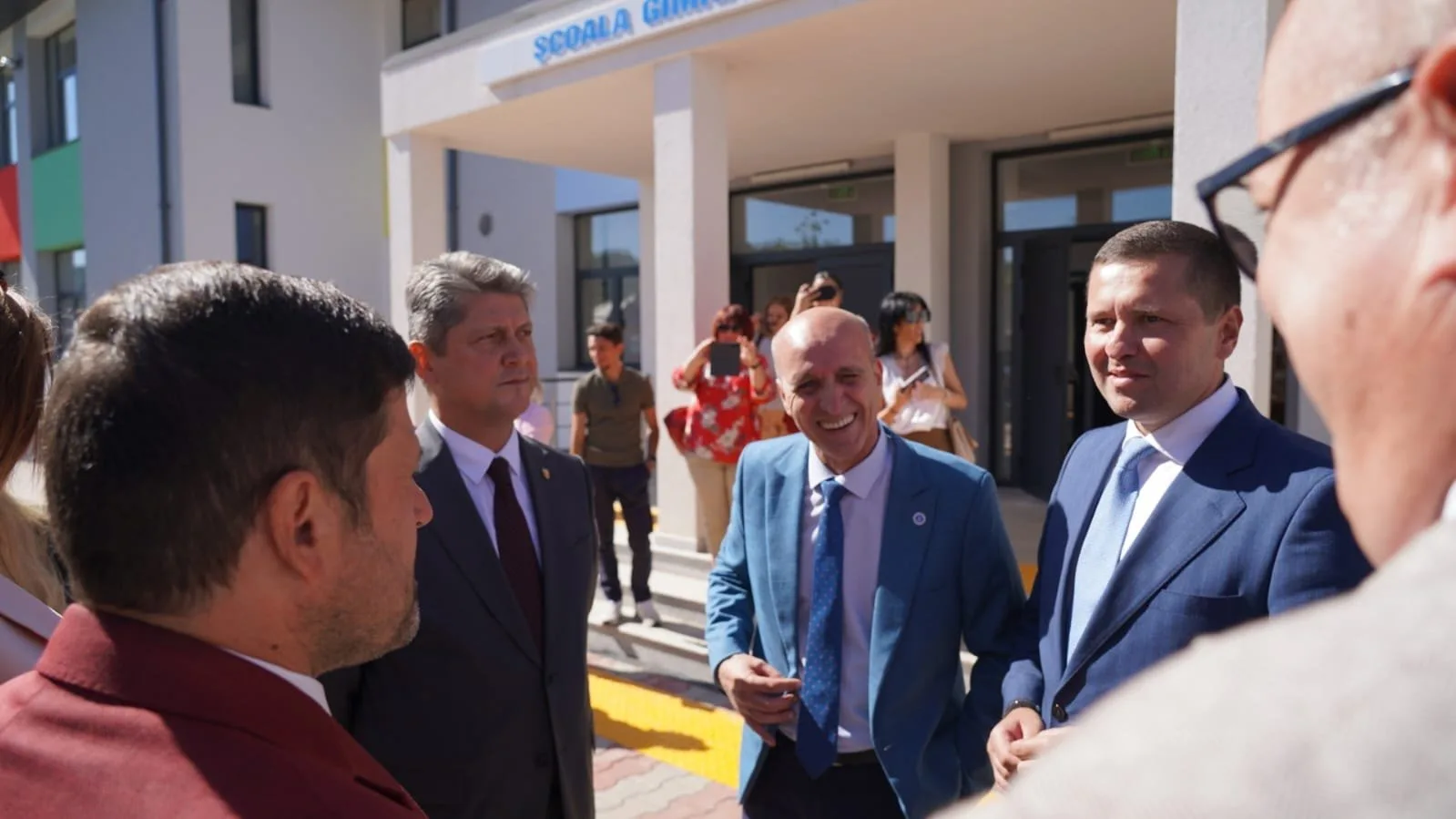 Titus Corlatean  and a group of men in suits engaged in conversation outside a building labeled 'Scuola G.' with several onlookers in the background.