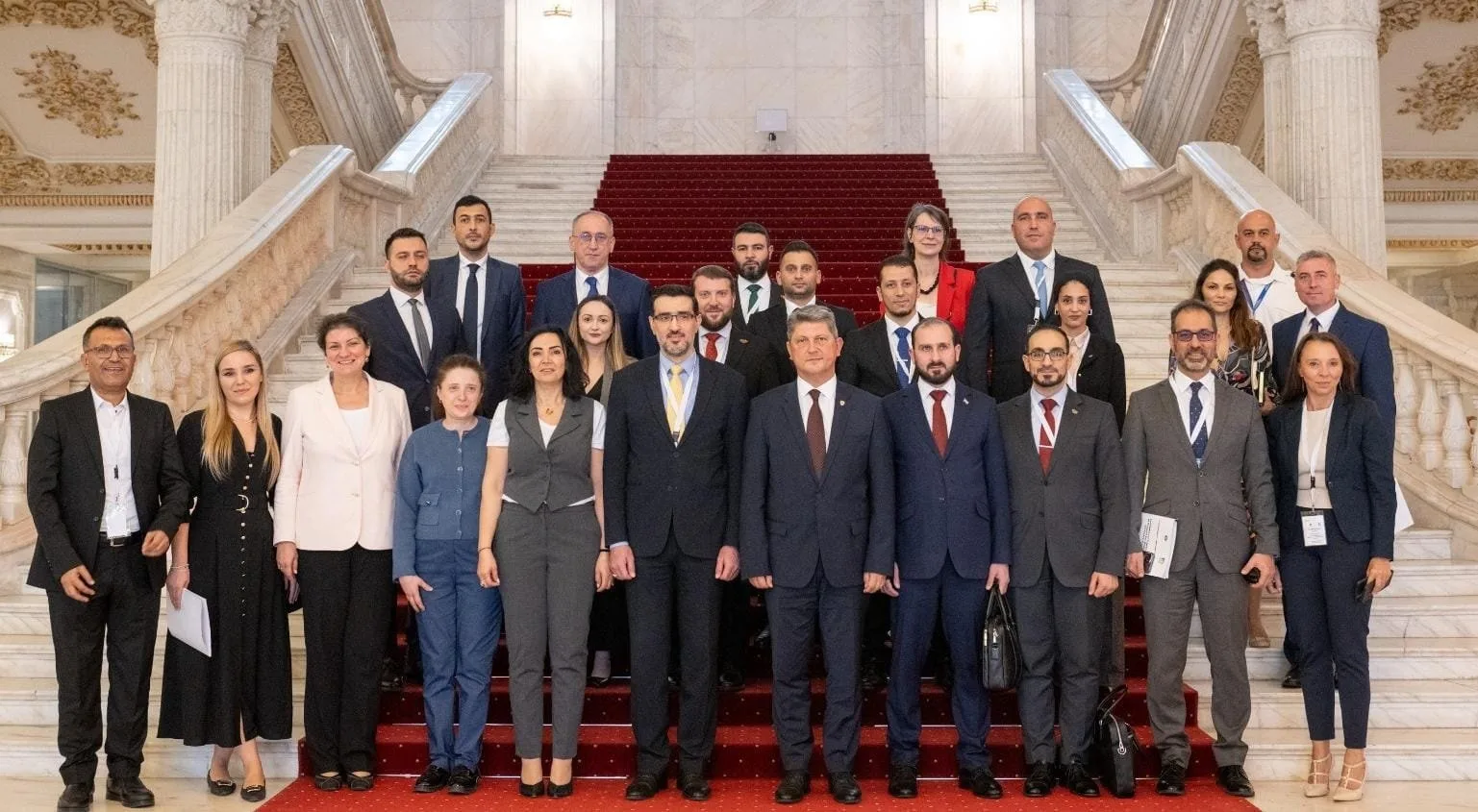Group of 21 people, men and women, standing on red-carpeted staircase inside a grand building with marble columns and ornate ceilings.