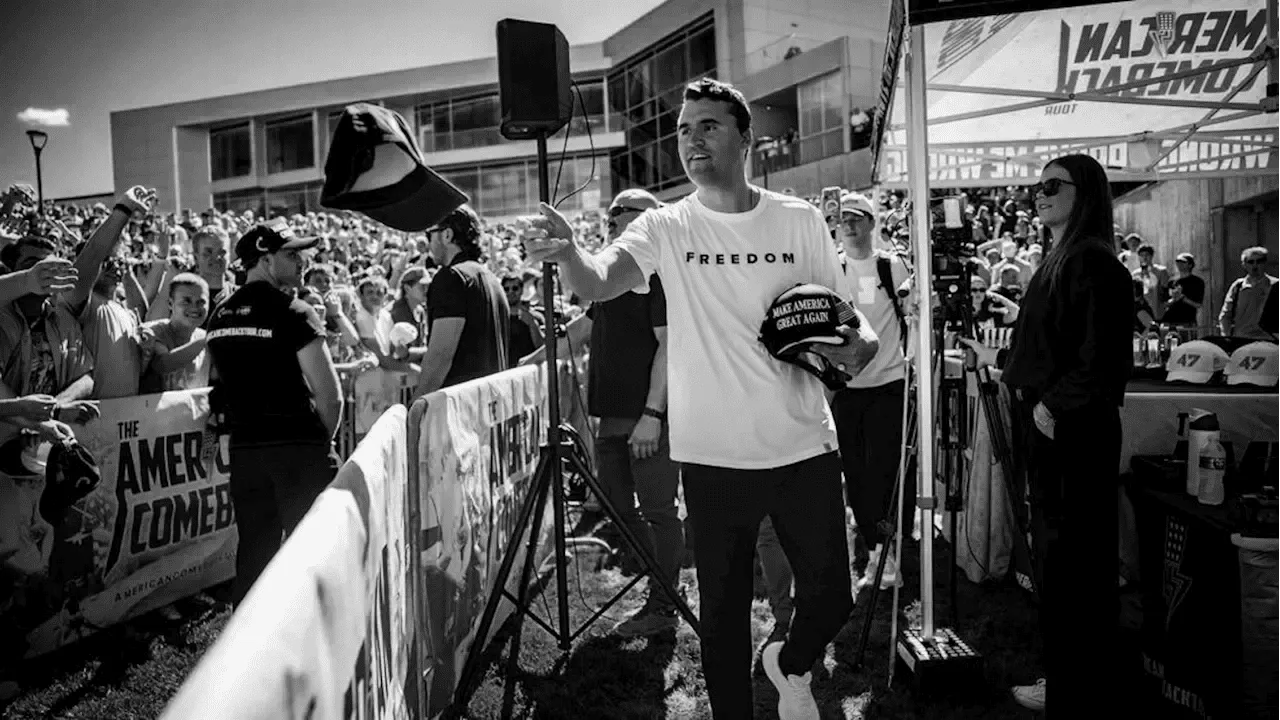 A man at a political rally or campaign event, wearing a T-shirt with 'FREEDOM' and holding a cap that says 'MAKE AMERICA GREAT AGAIN,' surrounded by a crowd and press.