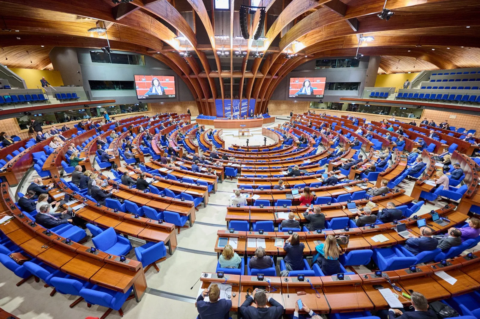 A large, modern conference chamber with people seated in blue chairs arranged in a semi-circular pattern around a central stage. The room features wooden interior design and multiple large screens displaying a presenter.