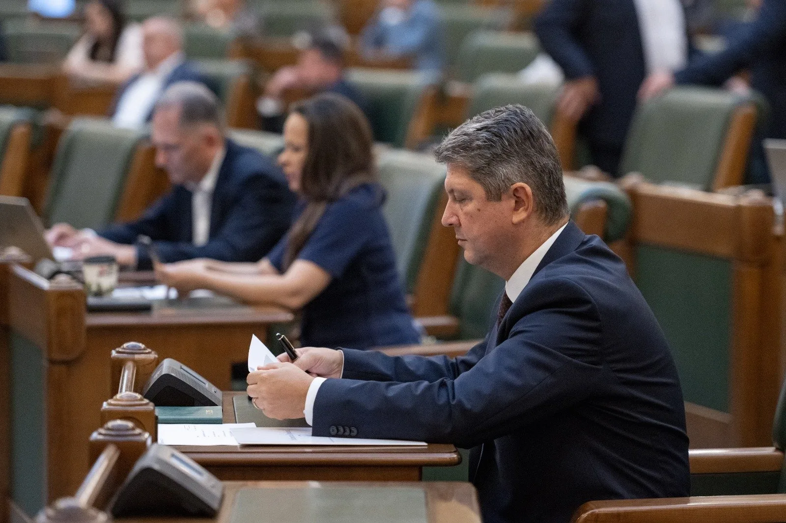 Titus Corlatean  sitting at a desk, looking at his phone in a large conference room filled with other people, some working on laptops, during a formal meeting or session.