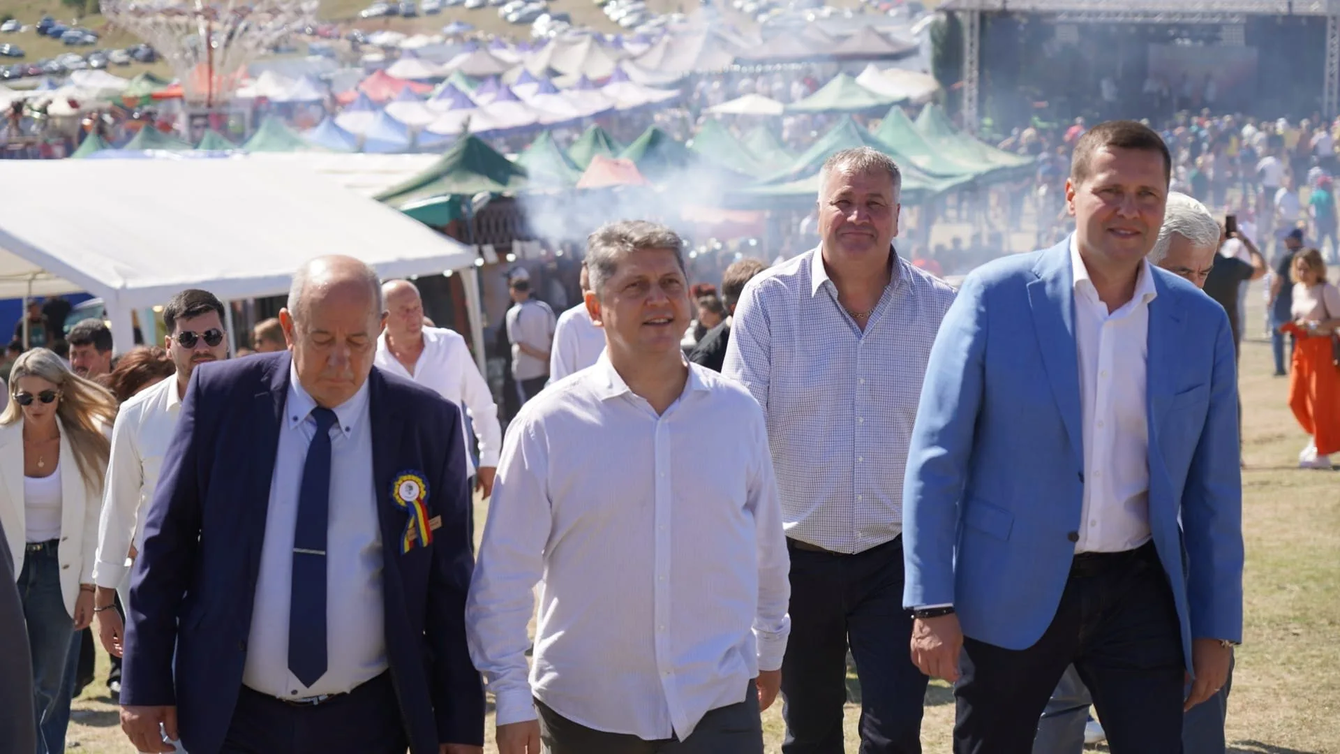 Group of four men walking at outdoor festival or fair with tents and people in the background.