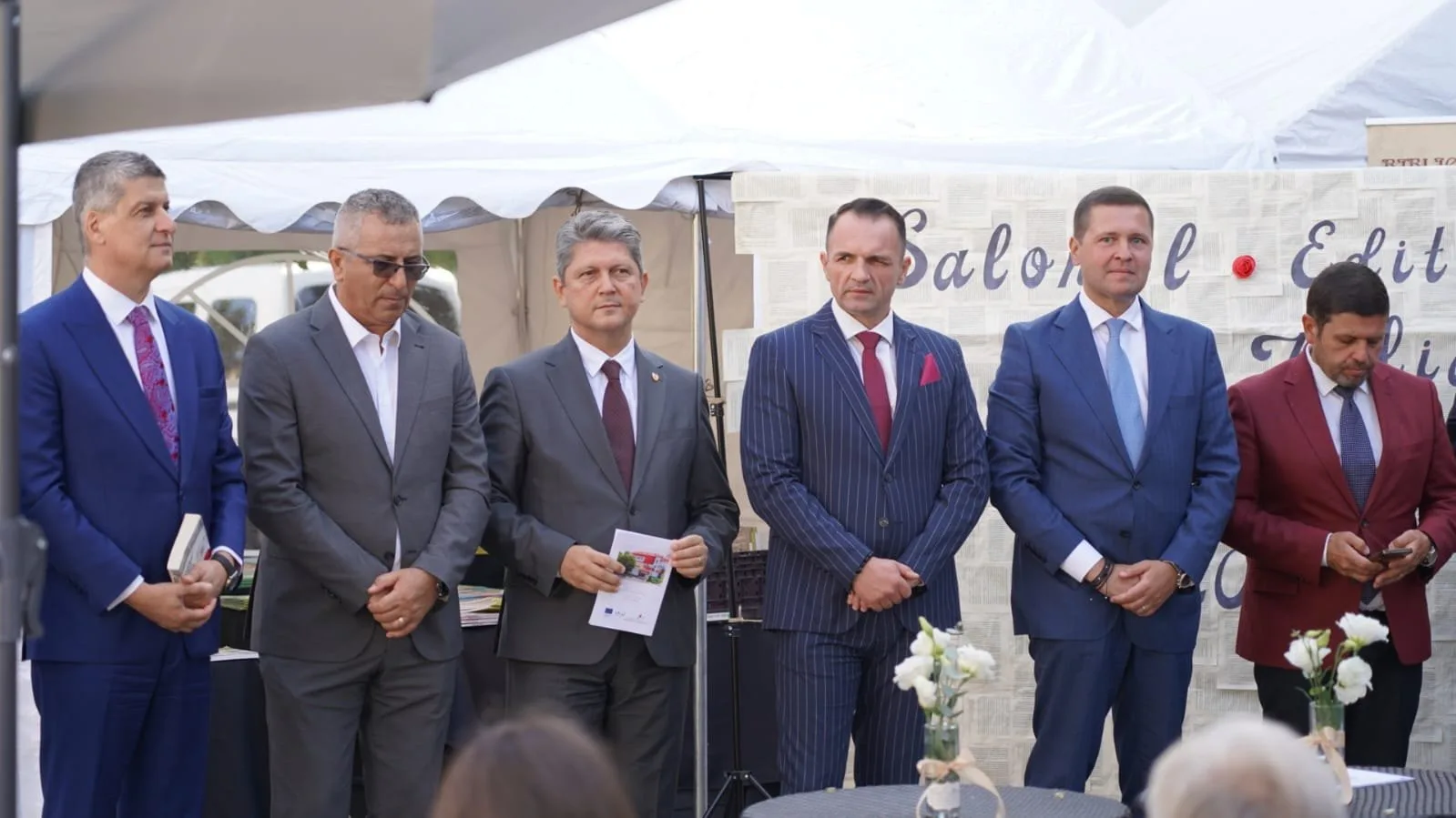 Titus Corlatean  and Six men in suits standing in a row at an outdoor event, under a white canopy tent, with a decorated backdrop and small tables with flowers in vases in front of them.