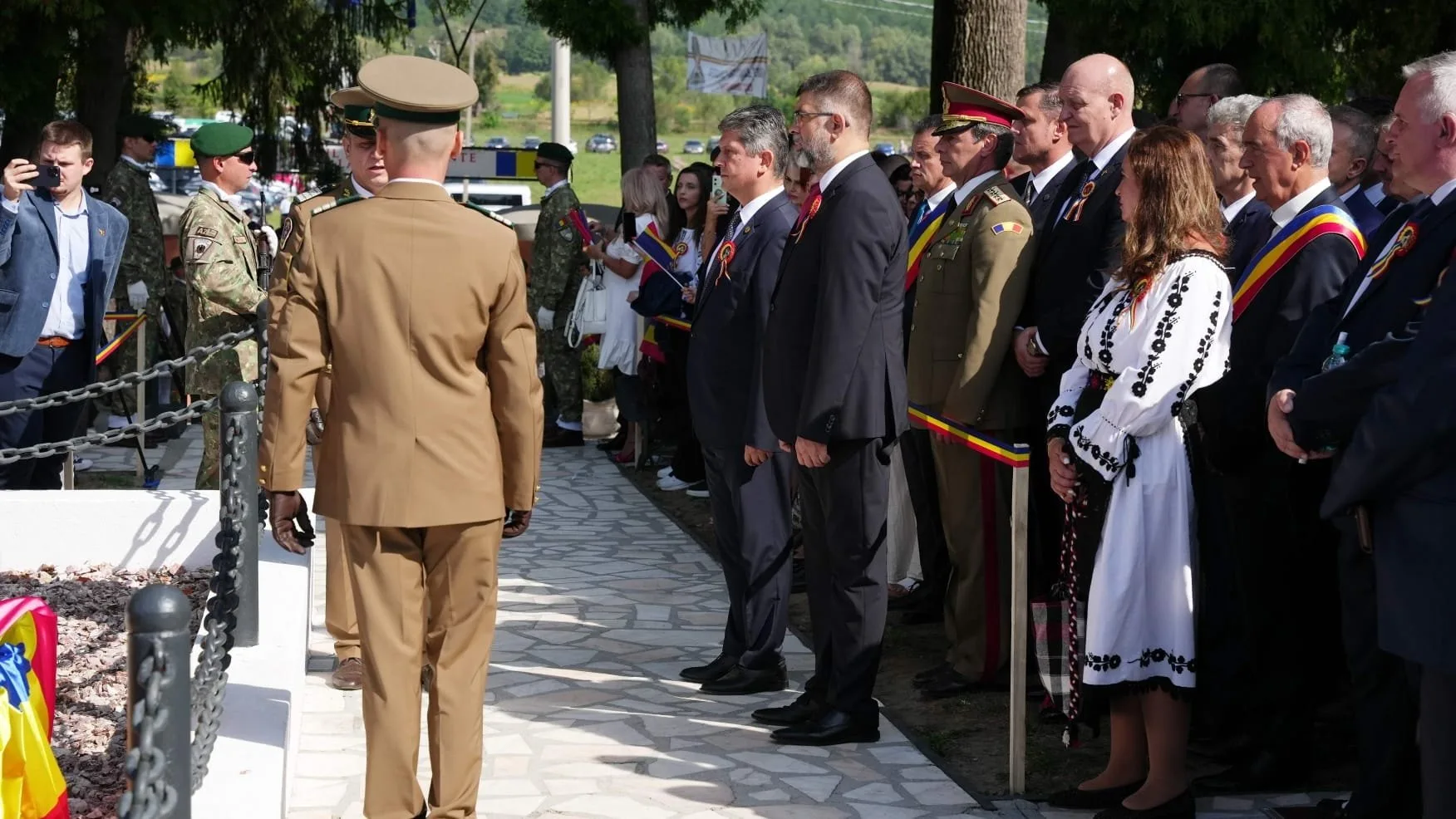 People gathered at a memorial or ceremony outdoors. Some are in military uniforms, and others are in formal attire, standing in a line with small Romanian flags. One person is taking a photo or video. The background includes trees and parked cars.