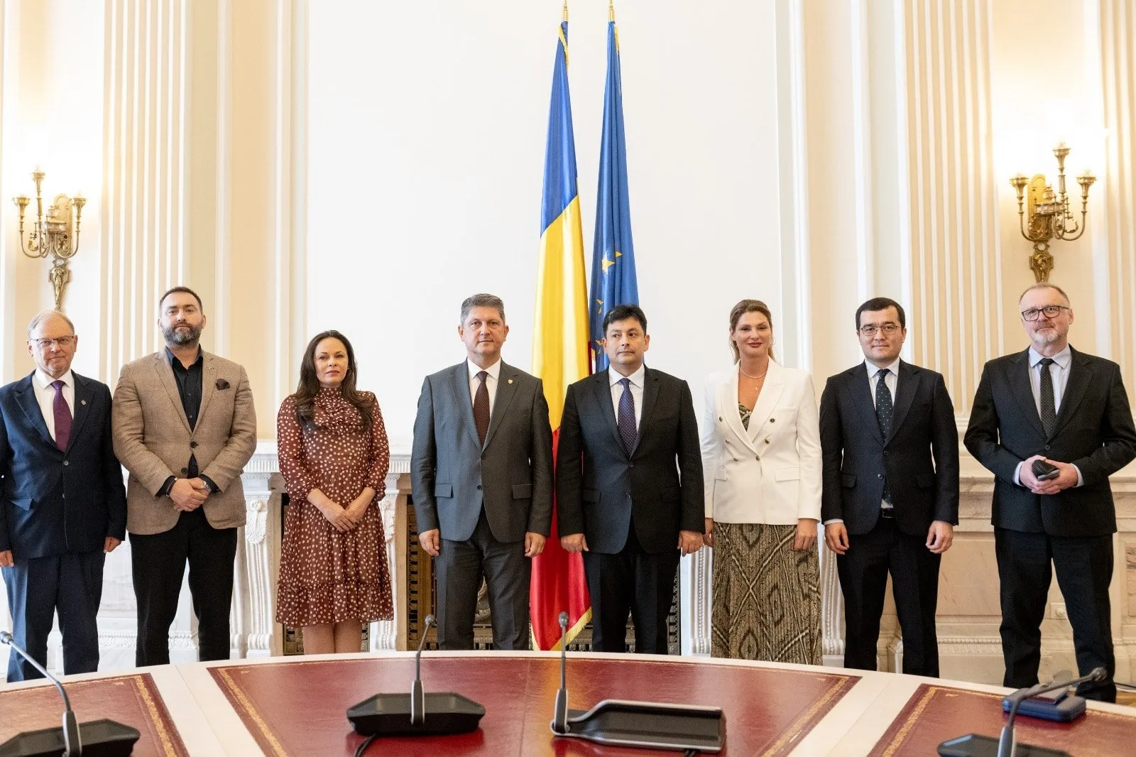 Group of nine people standing in front of national and European Union flags in a formal setting, with microphones on a conference table in the foreground.
